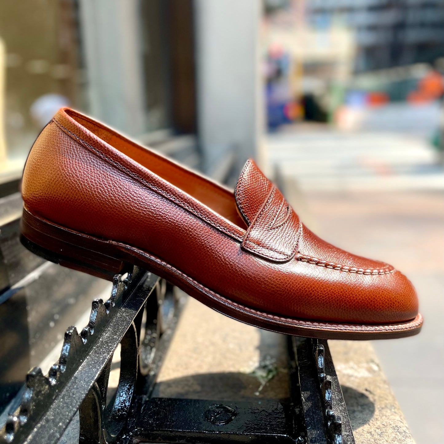 The Alden Madison D1204 - Alpine Grain NST Loafer in brown leather, featuring visible stitching and built on the Grant Last, is showcased outdoors on a black metal railing with a blurred urban background.