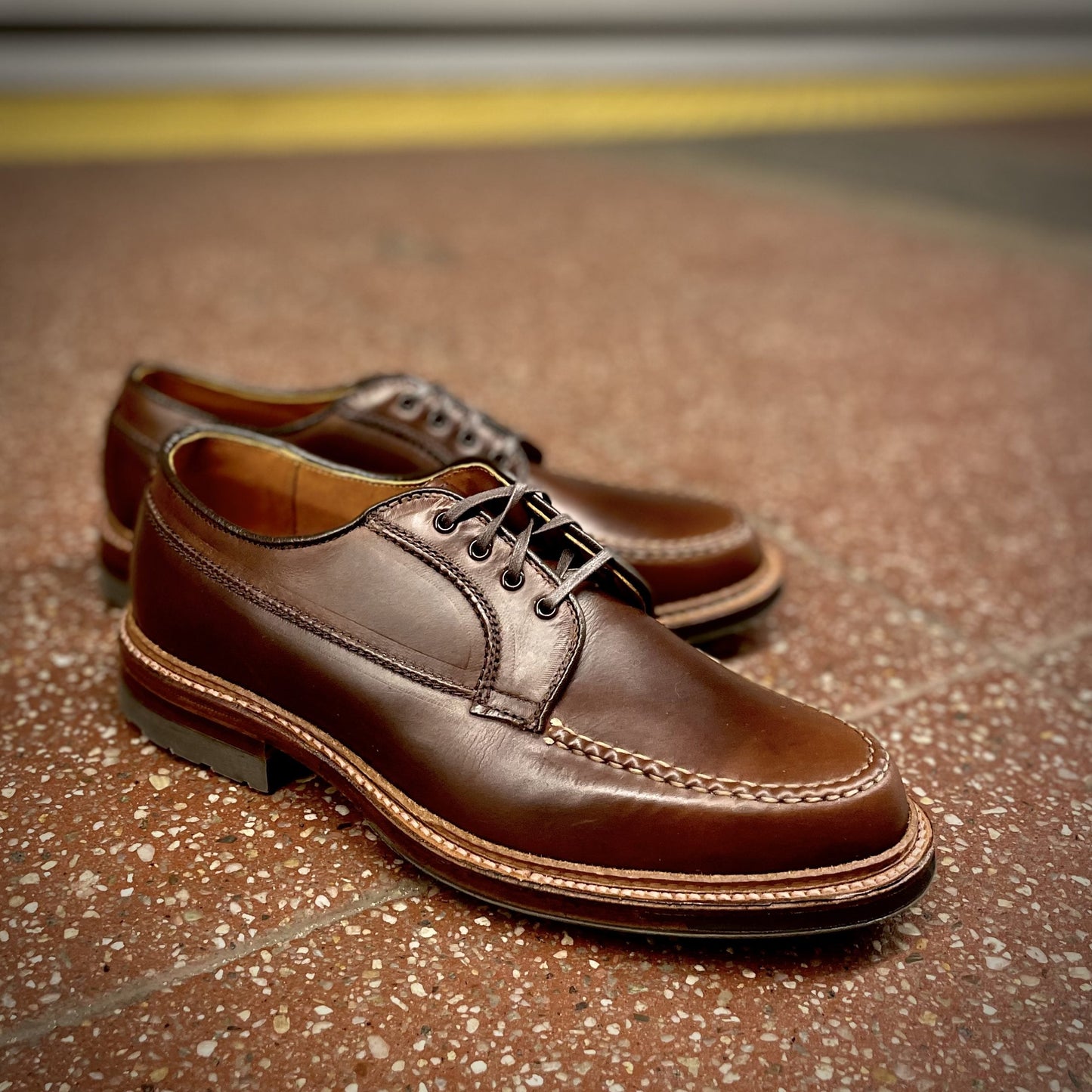 The Alden Madison D1611C - Ranger Mocc in Brown CXL features polished brown leather with detailed stitching, a slightly raised heel, and an Antique Edge for timeless elegance, all resting on a textured surface.