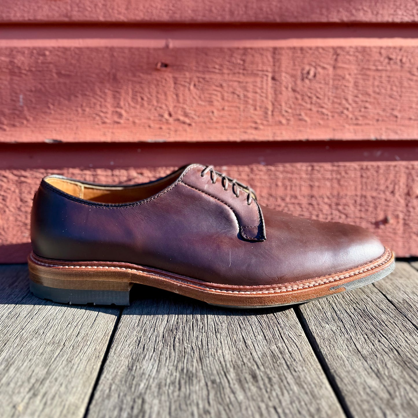 The ALDEN SHOES 9506C - Plain Toe Blucher w Commando Brown CXL, featuring black laces, is shown on a wooden surface before a red wooden wall, sunlight highlighting its brown Chromexcel leather.