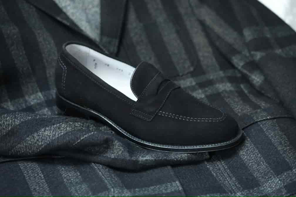 The Alden Madison D2229 “Stuyvesant” penny loafer in black suede with white stitching and a leather sole is showcased on a dark plaid-patterned suit jacket and matching scarf.