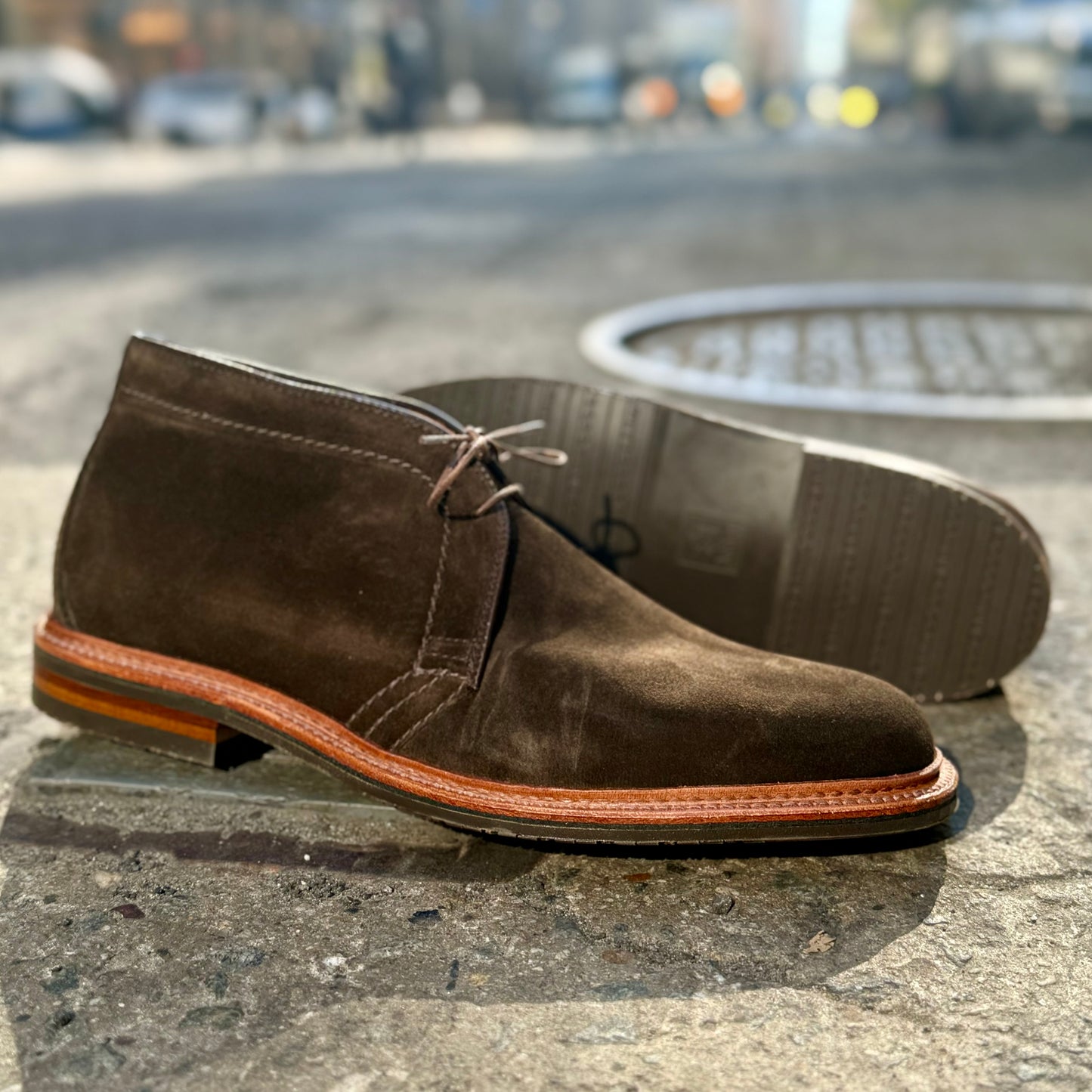 The D1712L Chukka Boot by Alden Madison, crafted on the Barrie Last in Loden Suede with a reverse welt, features a tan sole and laces on a sunlit sidewalk. Next to it, another boot lies on its side, highlighting its unique tread against a blurred urban backdrop.