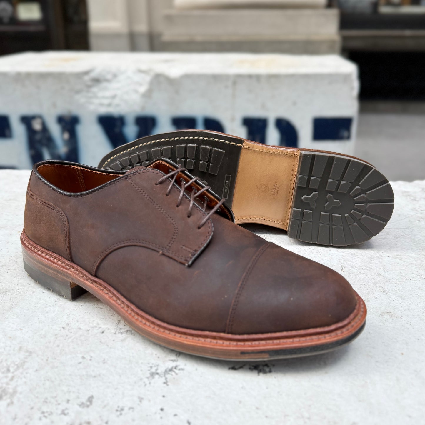 A pair of Alden Madison D4519C Tobacco Chamois Cap Toe Bluchers is on a concrete surface. The left shoe stands upright, highlighting the detailed lacing and cap toe stitching, while the right rests on its side, showing the textured sole and heel against a blurred urban backdrop.
