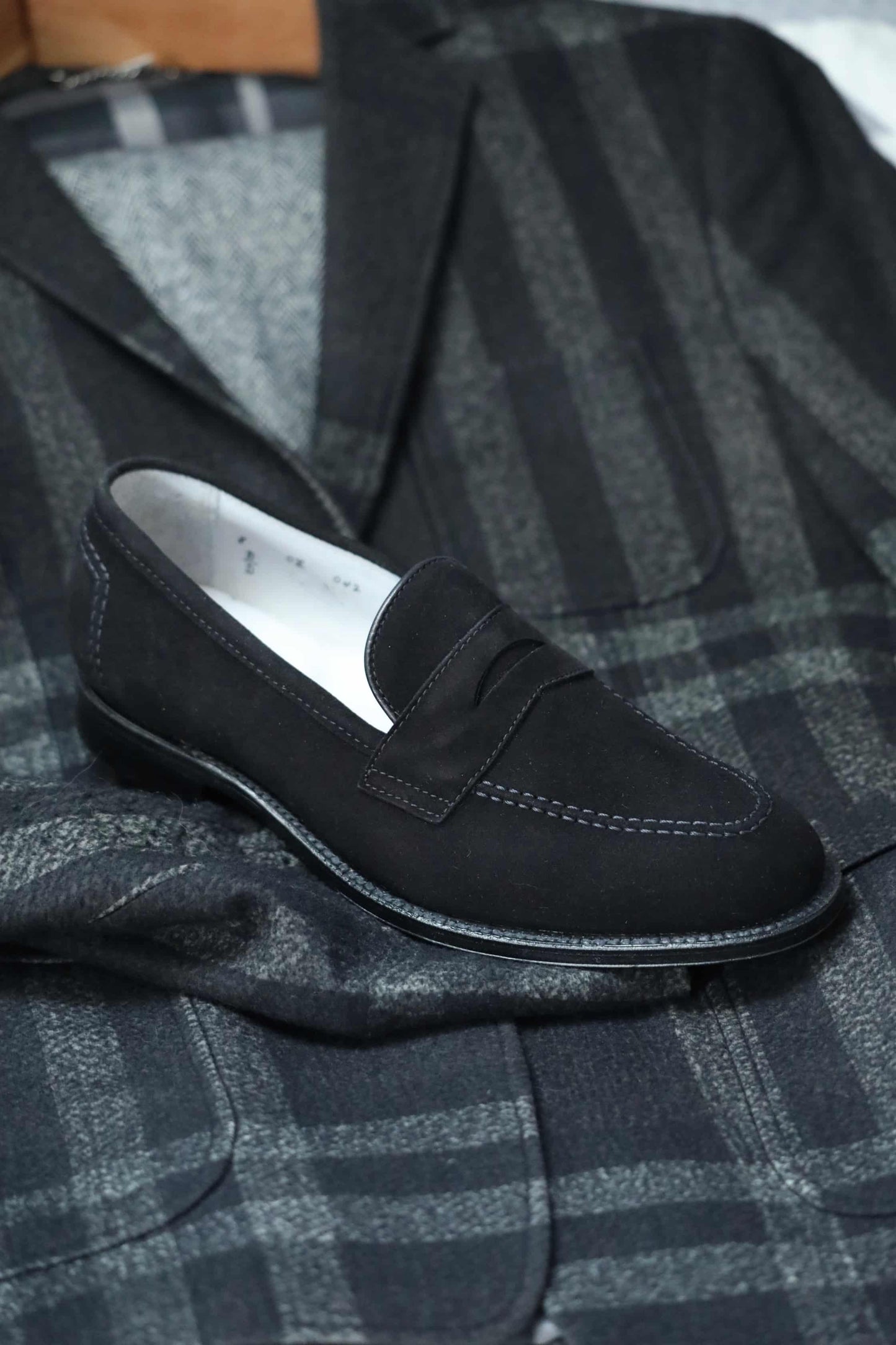 The Alden Madison D2229 “Stuyvesant” Madison Penny in Black Suede, featuring visible stitching and a timeless leather sole, sits on a dark plaid suit jacket for a classic, elegant look.