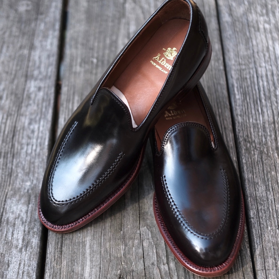 A pair of Alden Madisons D1118 - Vanderbilt Slip On in Color 8 Shell loafers, crafted from glossy dark brown Shell Cordovan leather with stitched details and a brand logo on the insole, rests on a weathered wooden surface.
