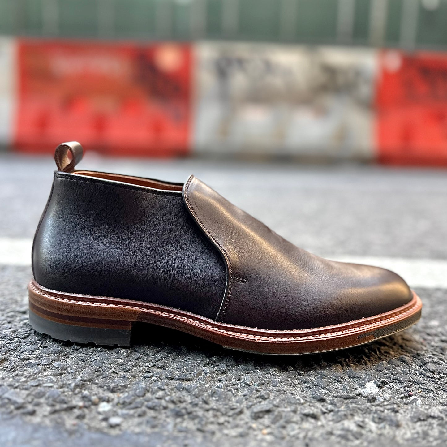 The Alden Madison D2714C - 750 Slip On Chukka in Arabica Lux, featuring a Commando rubber sole and pull tab, sits on an asphalt road with blurred red and white barriers in the background.