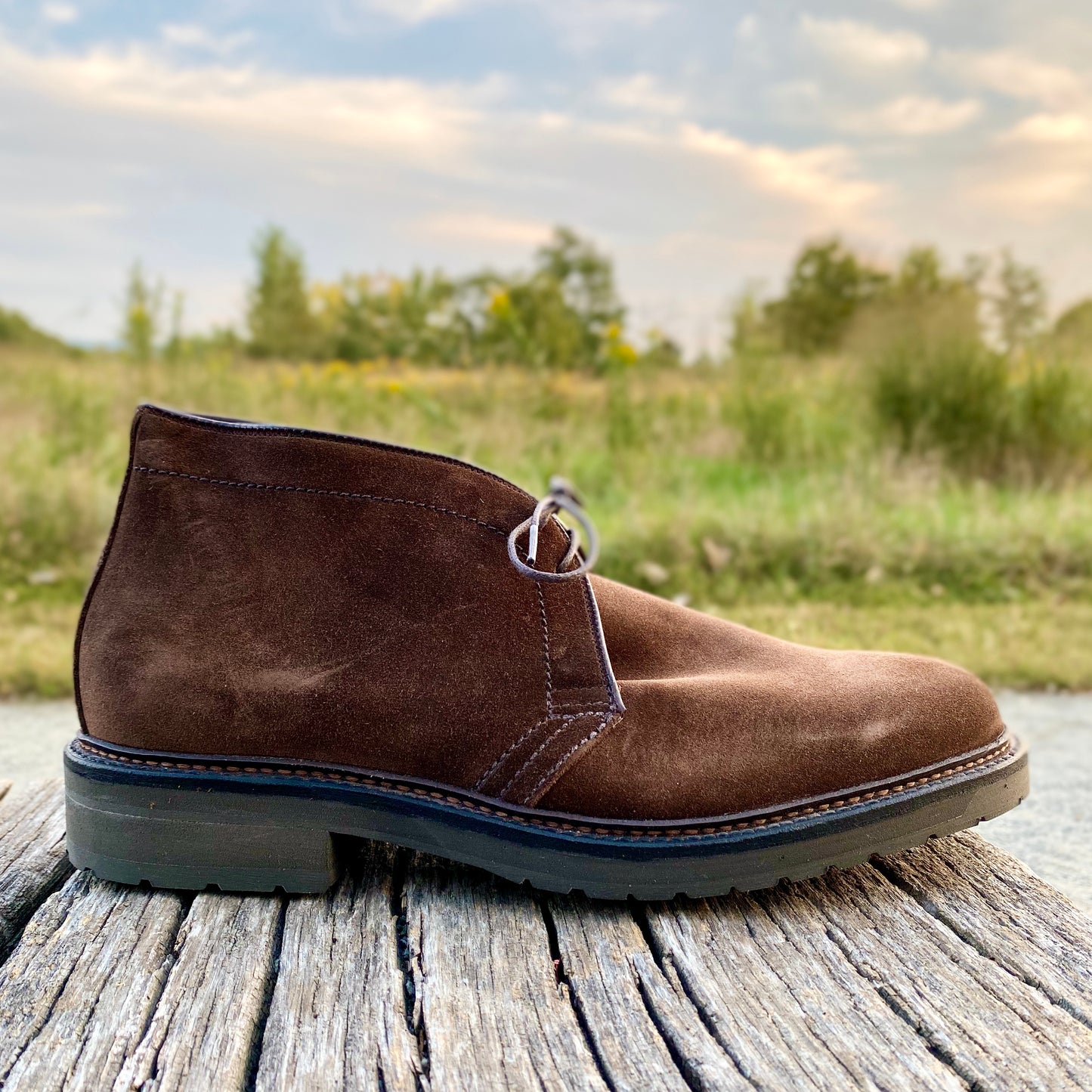 The Alden Madison 1273S - Chukka Boot in Brown Suede, featuring dark stitching and a thick lug rubber sole, rests on weathered wood with a grassy landscape and cloudy sky blurred in the background.