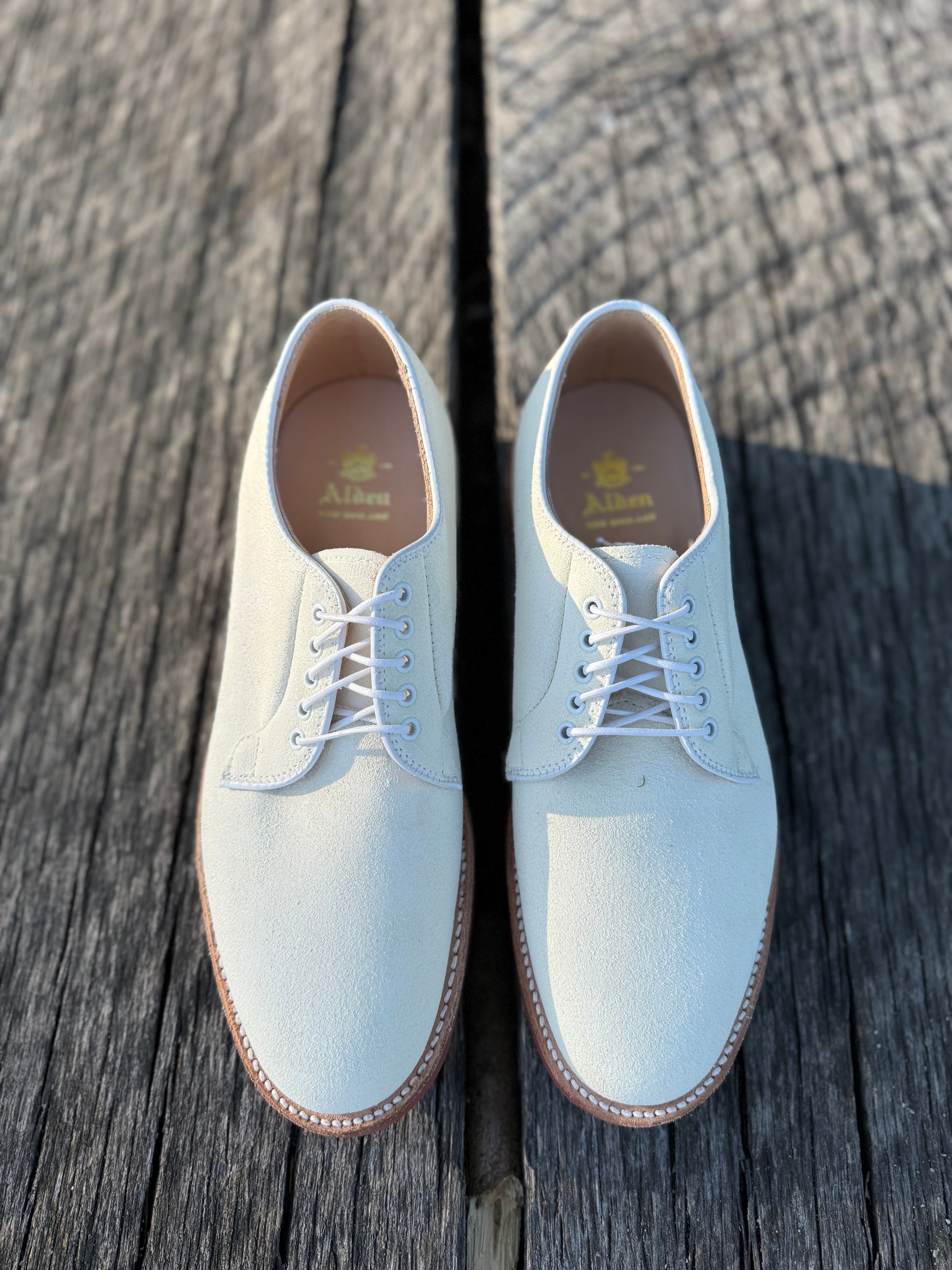 Alden Madison’s 9448 - White Buck in Ivory Suede derby shoes, crafted on the Barrie Last with brown soles, are displayed side by side on weathered wooden planks under bright sunlight.