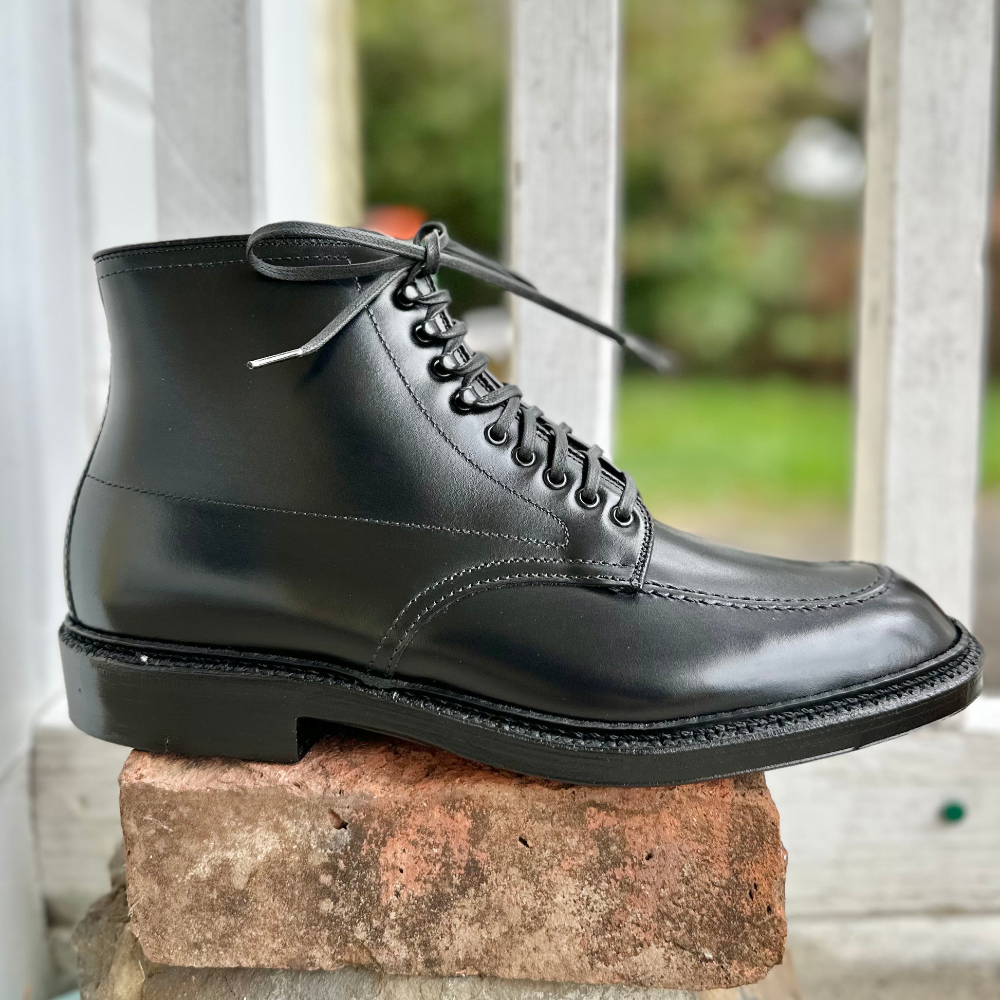 Displayed on a red brick, the Alden Madison D2941H - Black Calf Indy Boot showcases laces, fine stitching, and a double leather sole. Horizontal white railings and greenery form a beautifully blurred background.