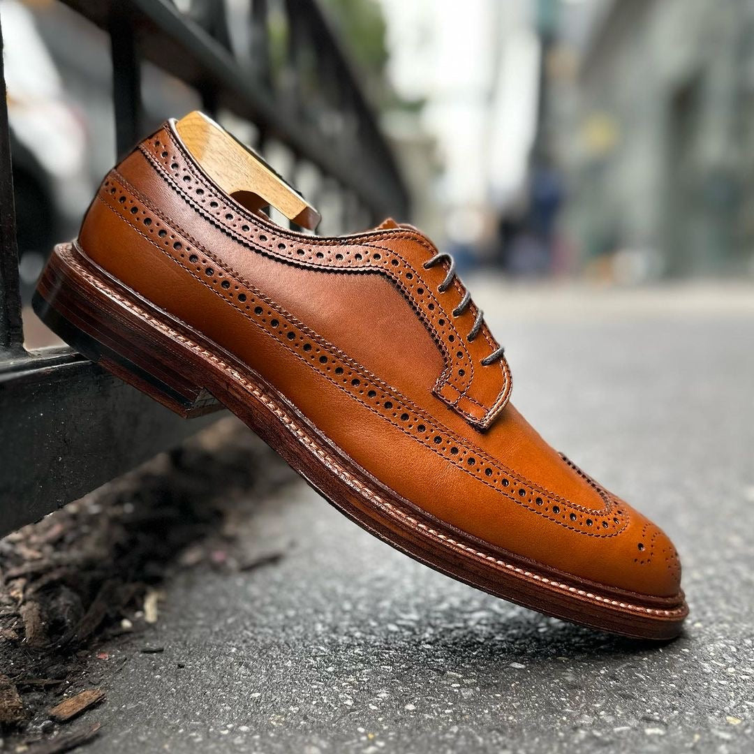 A brown burnished tan calfskin brogue from Alden Madisons 979 - Long Wing Blucher rests against a city sidewalk railing, featuring Barrie Last styles intricate perforations, sharply focused against a blurred urban backdrop.
