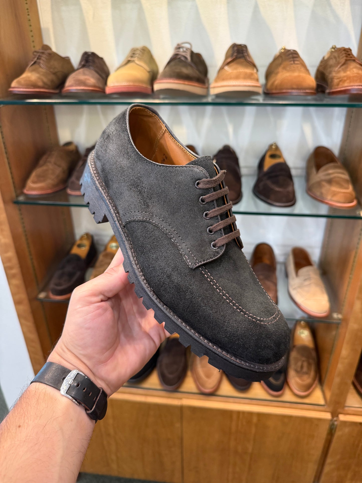 A hand holds the ALDEN SHOES 4006 - Earth Reverse Indy Shoe Heavy Lug, crafted on the Trubalance Last with a substantial lug rubber sole, in front of a display shelf lined with brown and tan dress shoes. The person is wearing a black watch.