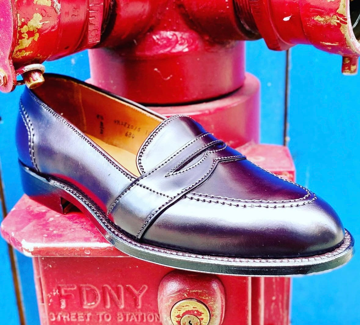 The Alden Madison 684 - Full Strap Loafer in Color 8 Shell Cordovan is showcased atop a red fire hydrant, set against a contrasting blue background.