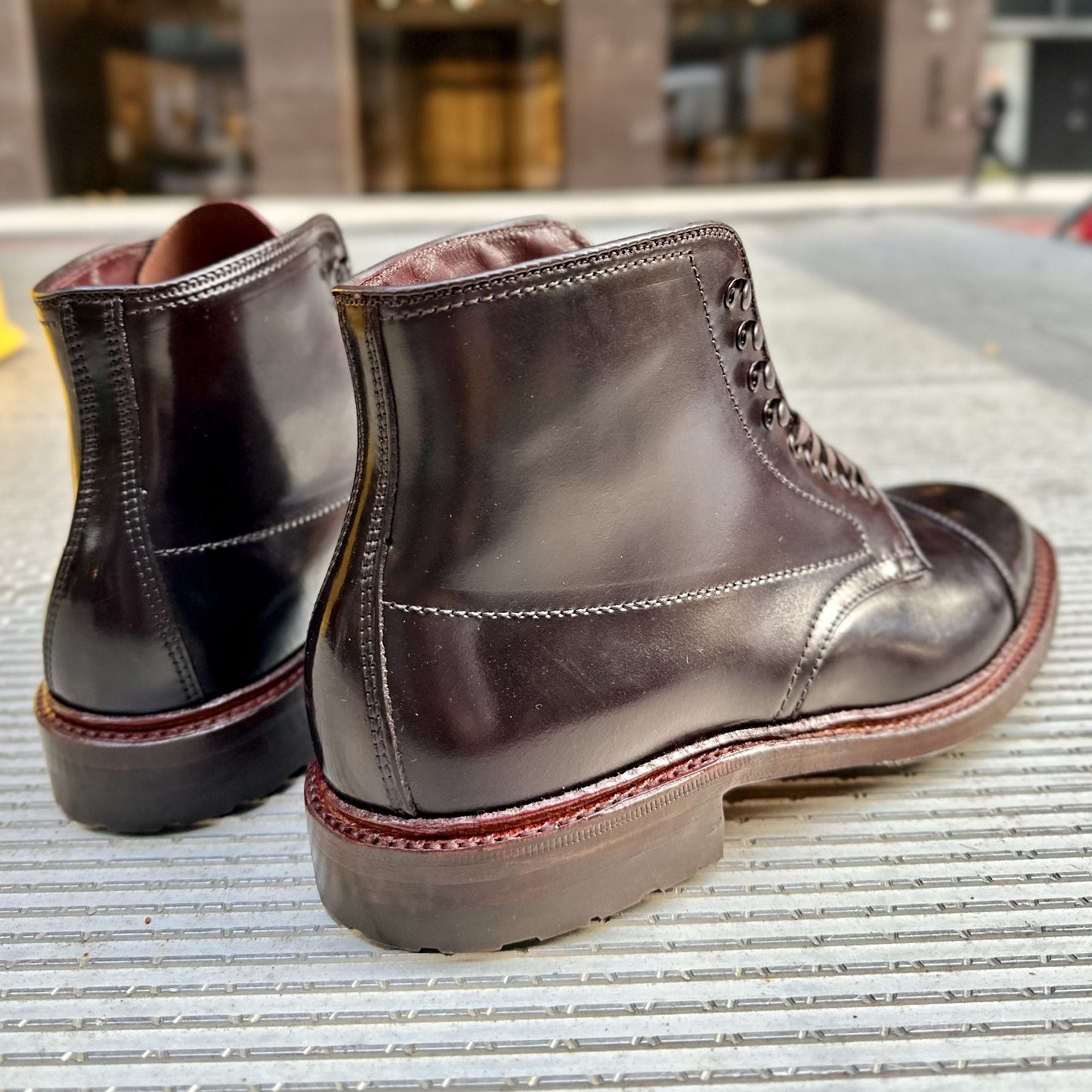 A close-up of sleek Alden Madison D2824HC Cap Toe Boots on a striped surface shows rich red-brown Color 8 Shell Cordovan soles with visible stitching, set against a blurred urban backdrop with towering buildings.