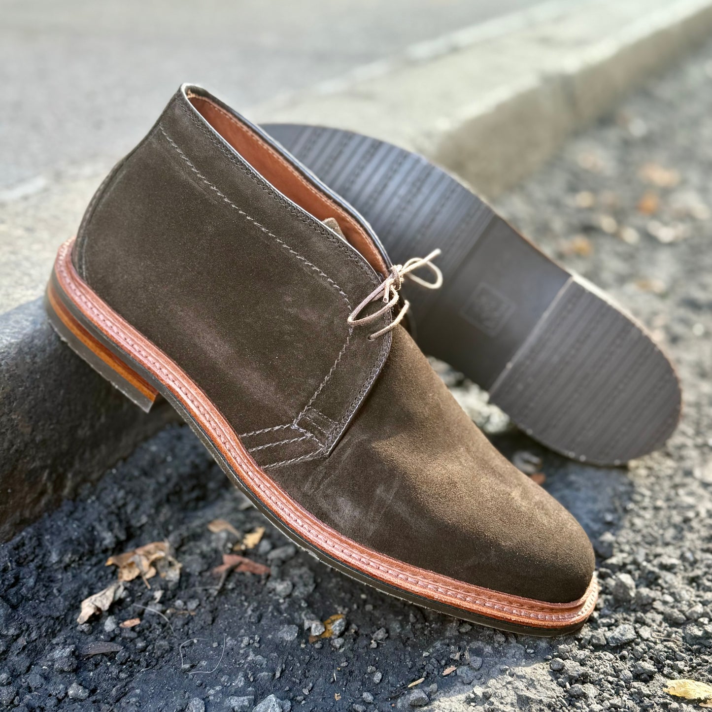 Aldens Madisons D1712L Chukka Boot in Loden Suede, featuring an L3 sole and reverse welt, sits on a concrete curb. One stands upright, the other lies on its side, highlighting the sole against a gravelly backdrop.