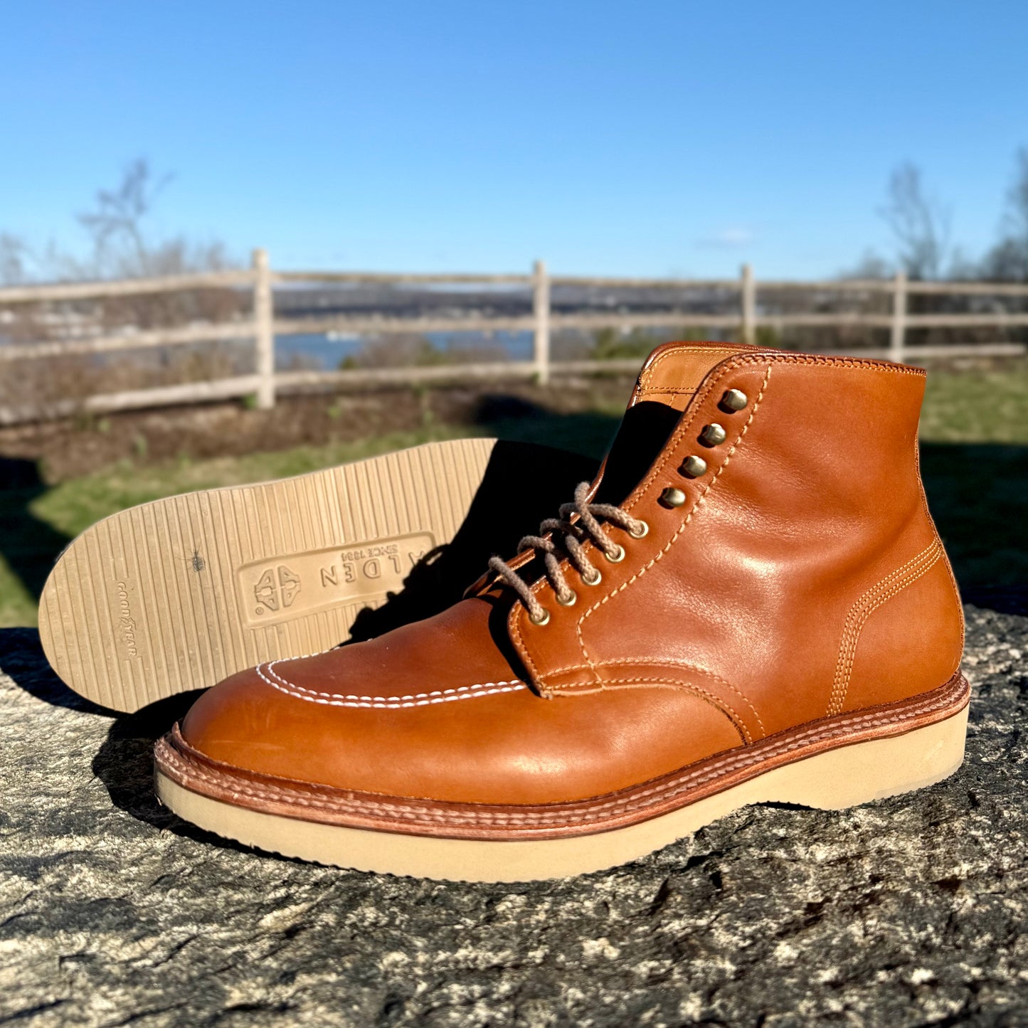 A pair of 40311H - Sherwood Indy Boots by Alden Madison, made from Saddle Calf leather with light soles, is shown on a stone surface outdoors. One boot stands upright while the other lies on its side, showing the sole, with a wooden fence and lake blurred in the background.