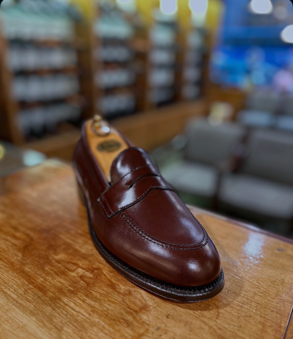 The Alden Madison D5214 - Brown Calf Flex Penny loafer, expertly crafted in rich brown calf leather on the Copley Last and finished with a flexible leather sole, is showcased on a wooden surface against a softly blurred backdrop of shelves and seating inside a shoe store.