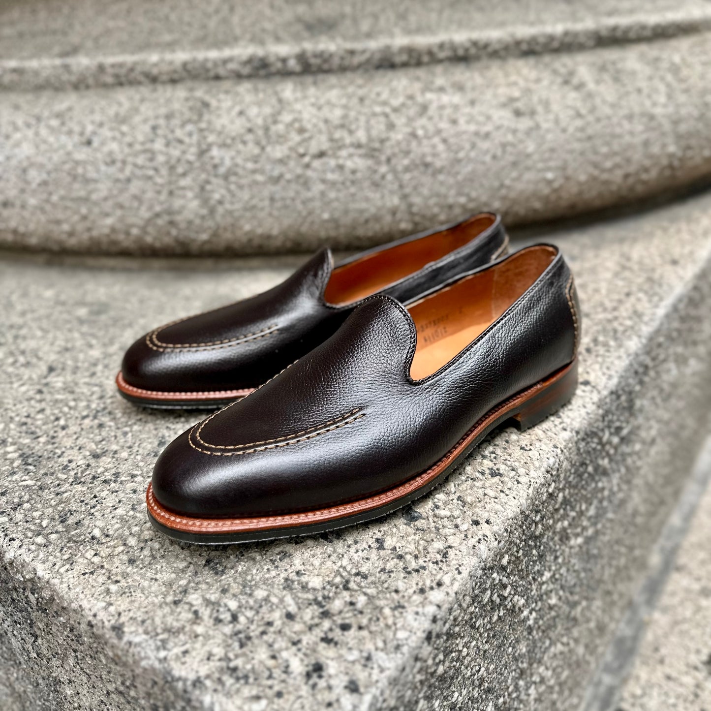 Alden Madison’s A1101L - Vanderbilt in Brown Regina Grain loafers feature dark brown grain calfskin, light stitching, a tan interior, and a durable rubber sole, shown on grey stone steps.