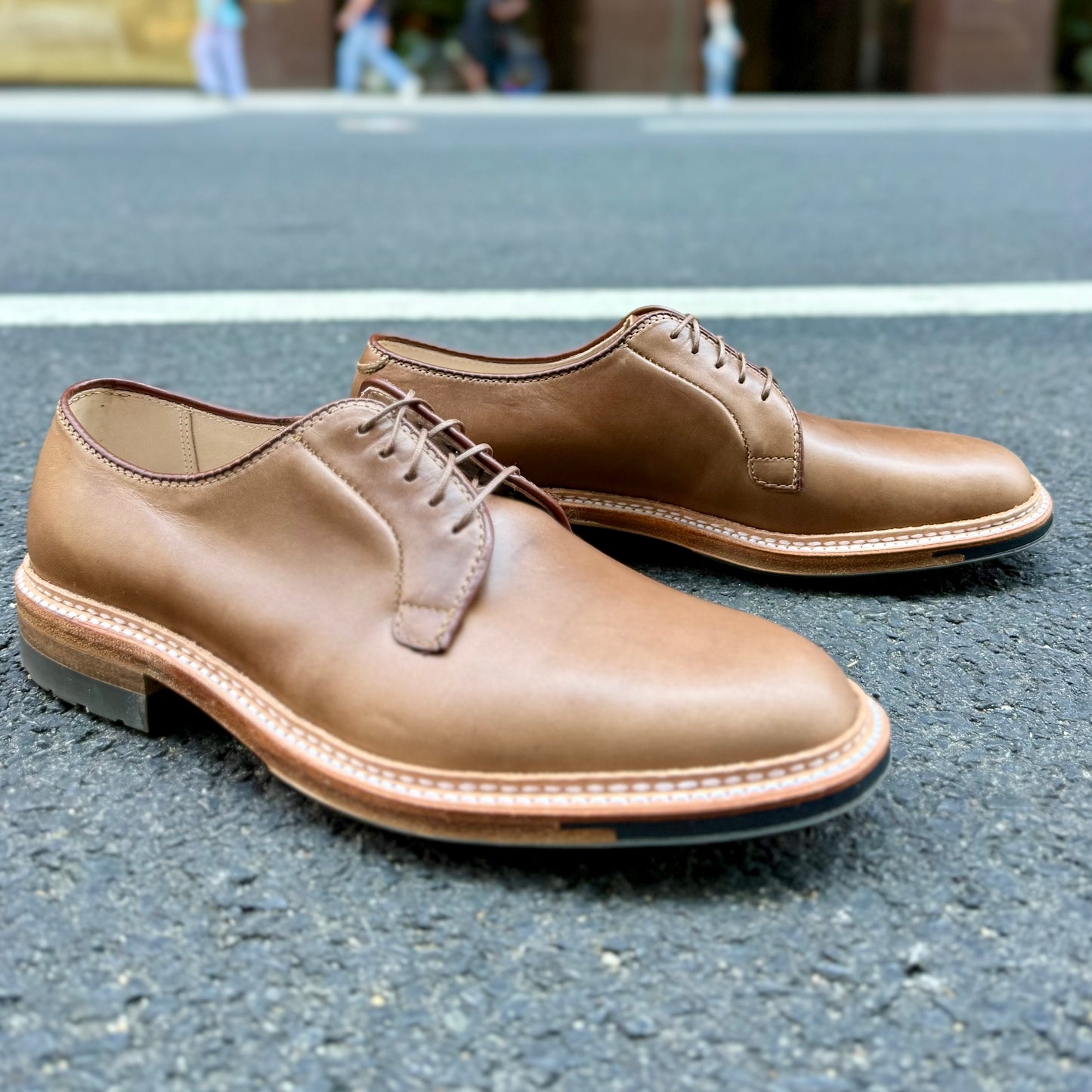 A pair of ALDEN SHOES 9501C - Natural CXL Plain Toe Bluchers with laces rests on an asphalt road, with a blurred street and people in the background.