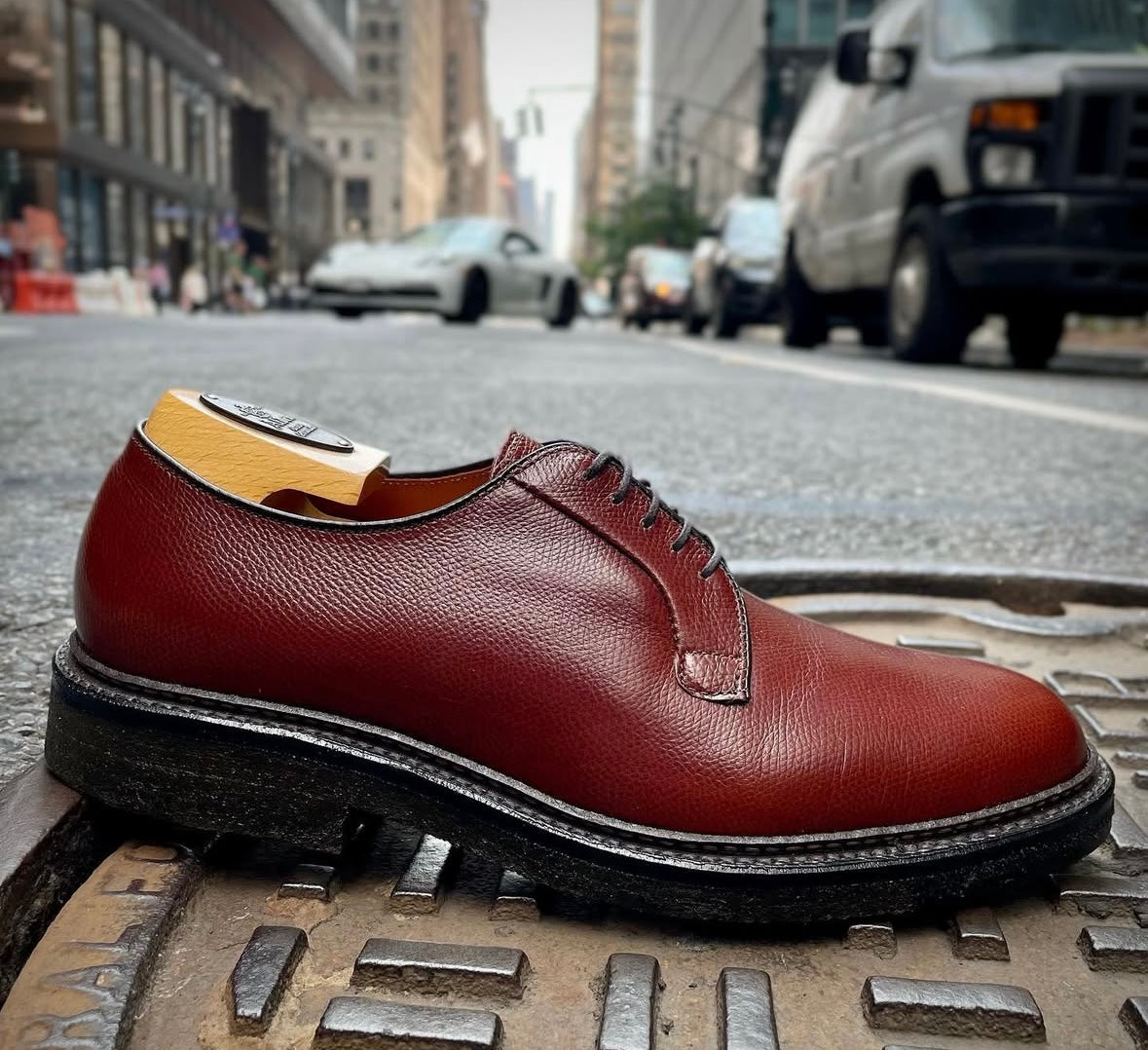 A single Alden Madison 947 - All Weather Walker Brown Alpine Grain dress shoe with black laces and a crepe sole sits on a manhole cover in the city street, surrounded by blurred vehicles and urban buildings.