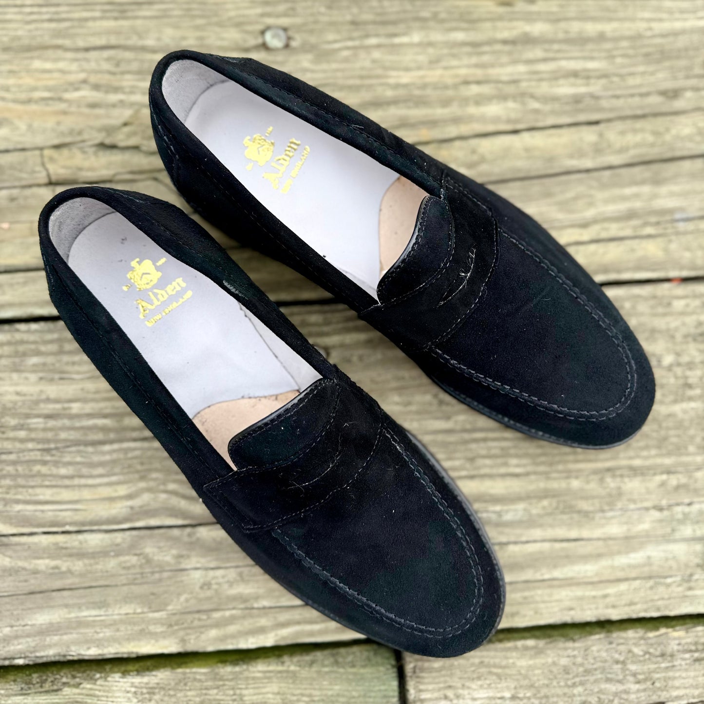 A pair of Alden Madison D2229 “Stuyvesant” Madison Penny loafers in black suede, featuring gold insole branding and leather soles, displayed on weathered wooden planks.