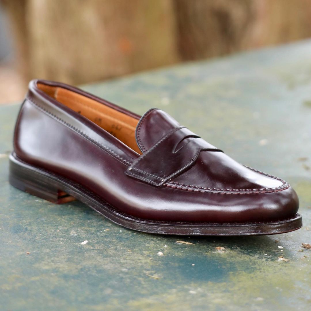 Alden Madisons 986 - Leisure Hand Sewn loafer in Color 8 Shell Cordovan is showcased on a textured green surface. This polished brown leather shoe features a sleek design with low heel, visible stitching, and penny strap against a blurred background, highlighting its artisan craftsmanship.