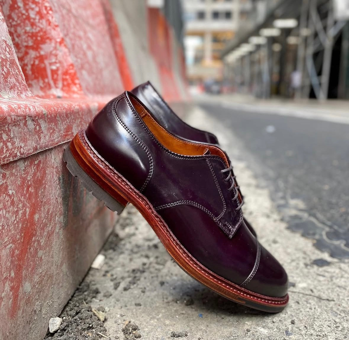 A pair of Alden Madison D8516C DeRosa Captoe Blucher shoes in Color 8 Shell, featuring an Antique Edge and brown soles, rests against a red and white city street barrier.