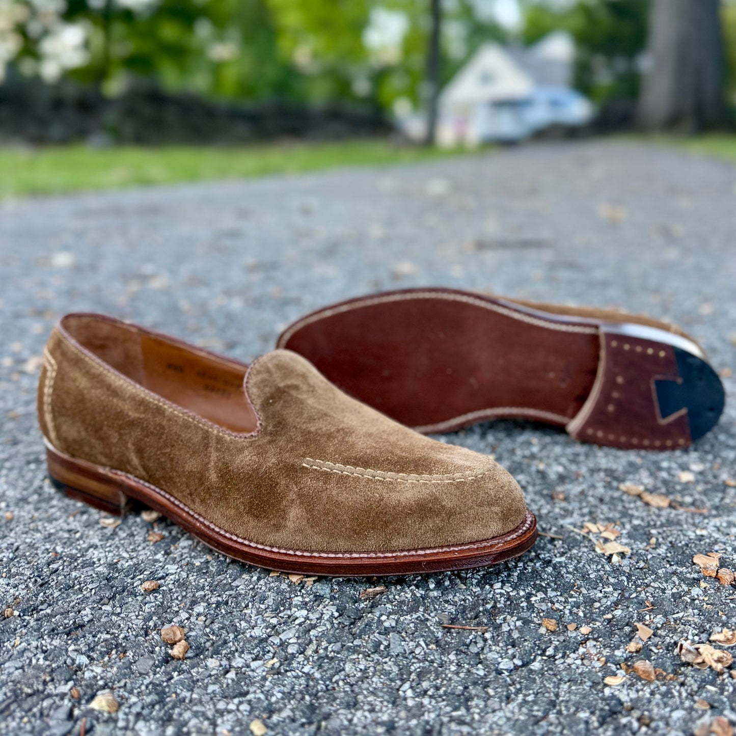 A pair of Alden Madison D2101 - Vanderbilt Slip On in snuff suede sit on the pavement, crafted with sturdy leather soles; one shoe stands upright, the other lies on its side with greenery and a house blurred in the background.