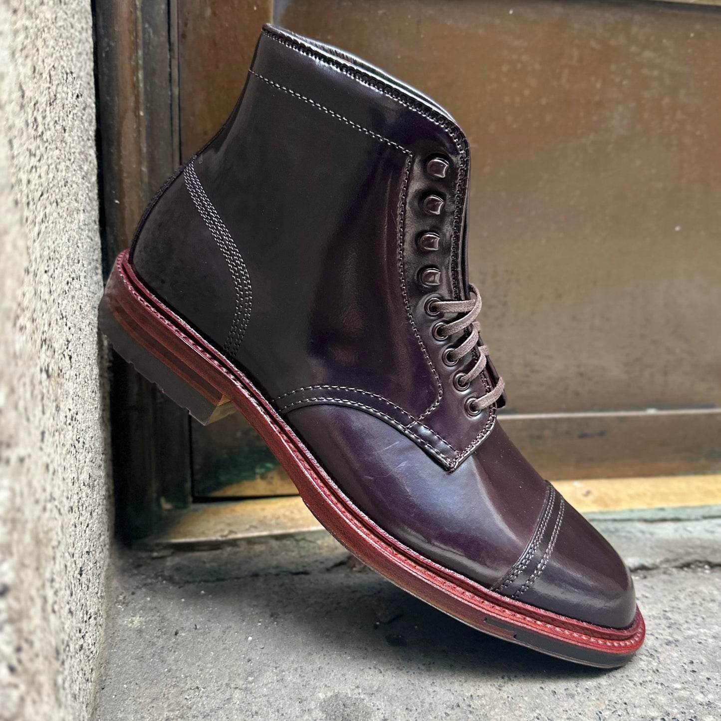 Alden Madisons D2858HC - 2/2 Cap Boot in Color 8 Shell Cordovan, featuring a polished dark brown finish, detailed stitching, lacing eyelets, and a signature red commando sole, stands elegantly against a rough concrete surface.