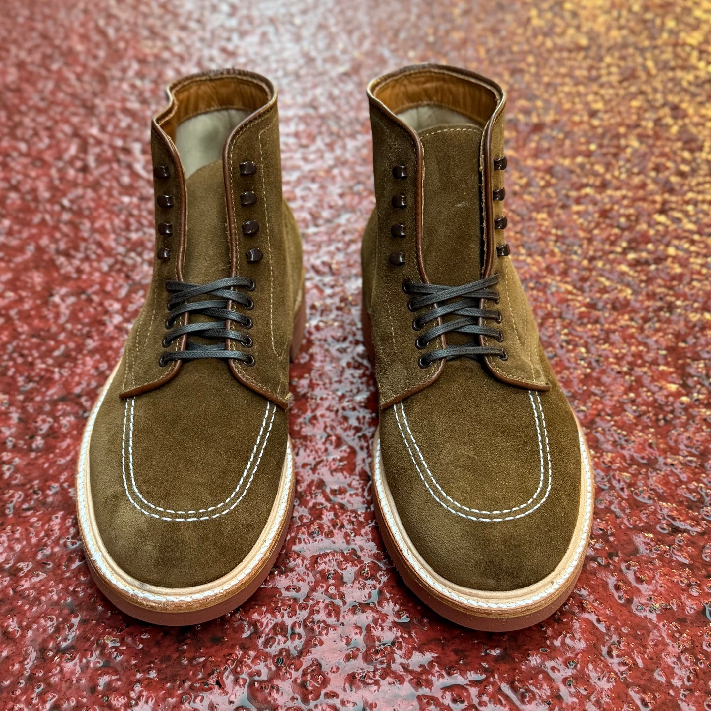 The 40531H - Redwood Indy Boot in Snuff Suede by Alden Madison features black laces and visible white stitching, displayed on a textured red surface.