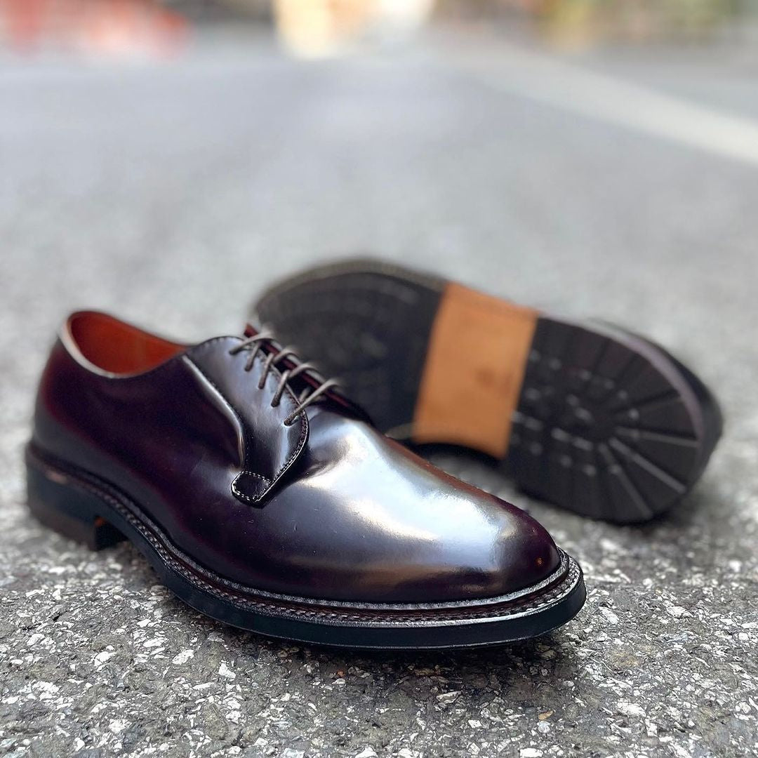 The Alden Madison 990C Plain Toe Blucher in Color 8 is showcased on a gray surface, with one dress shoe upright and its partner on its side, emphasizing the shell cordovan leather and durable commando sole craftsmanship.