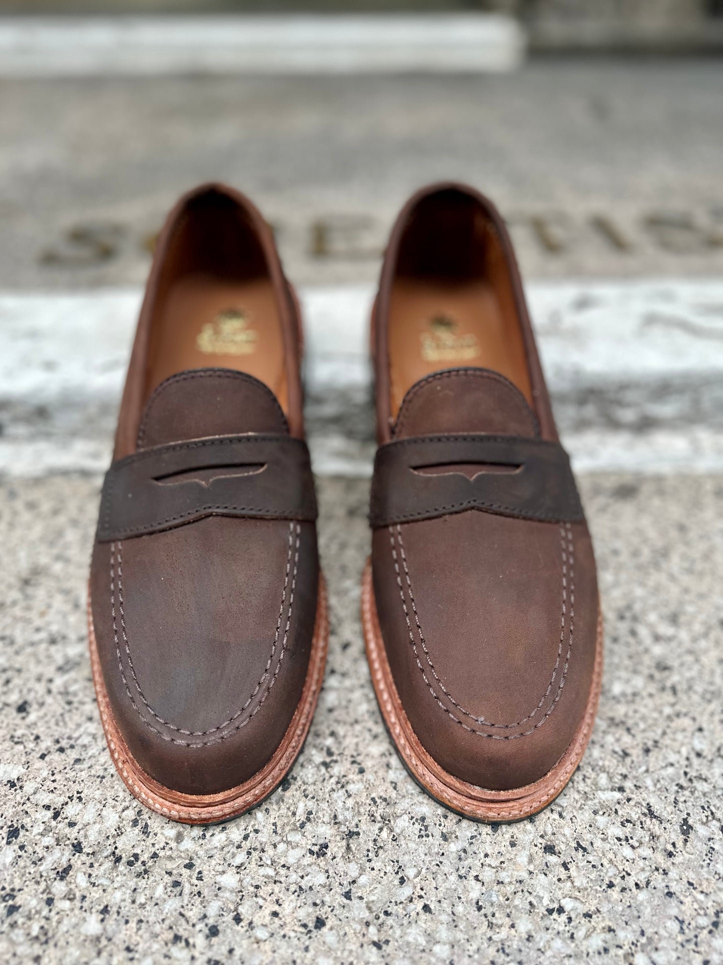 Alden Madisons D2216L - Smooth Tobacco Chamois Penny Loafers, with stitching details and a small strap cutout, rest on a textured gray surface. The blurred background highlights their antique edge and rich finish.