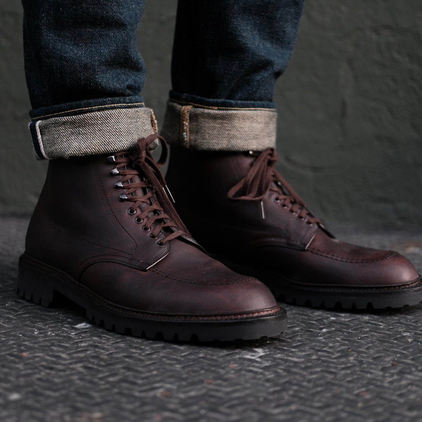 A person is wearing Alden Madisons 404 - Indy Boot in Kudu (Oiled CXL), polished dark brown lace-up boots with a durable Vibram Lug Outsole. Theyre paired with cuffed dark blue jeans and resting on a textured metal surface against a muted green wall.