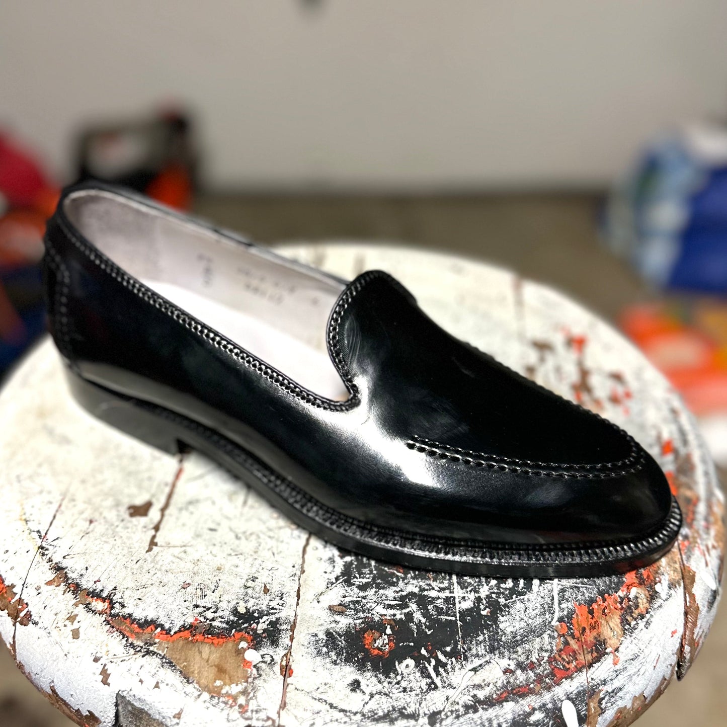 A single, elegant Vanderbilt Slip On in Black Shell Cordovan by Alden Madison with stitching details and a classic Brooks heel rests on a worn, painted wooden stool. The blurred background emphasizes the loafers sophistication.