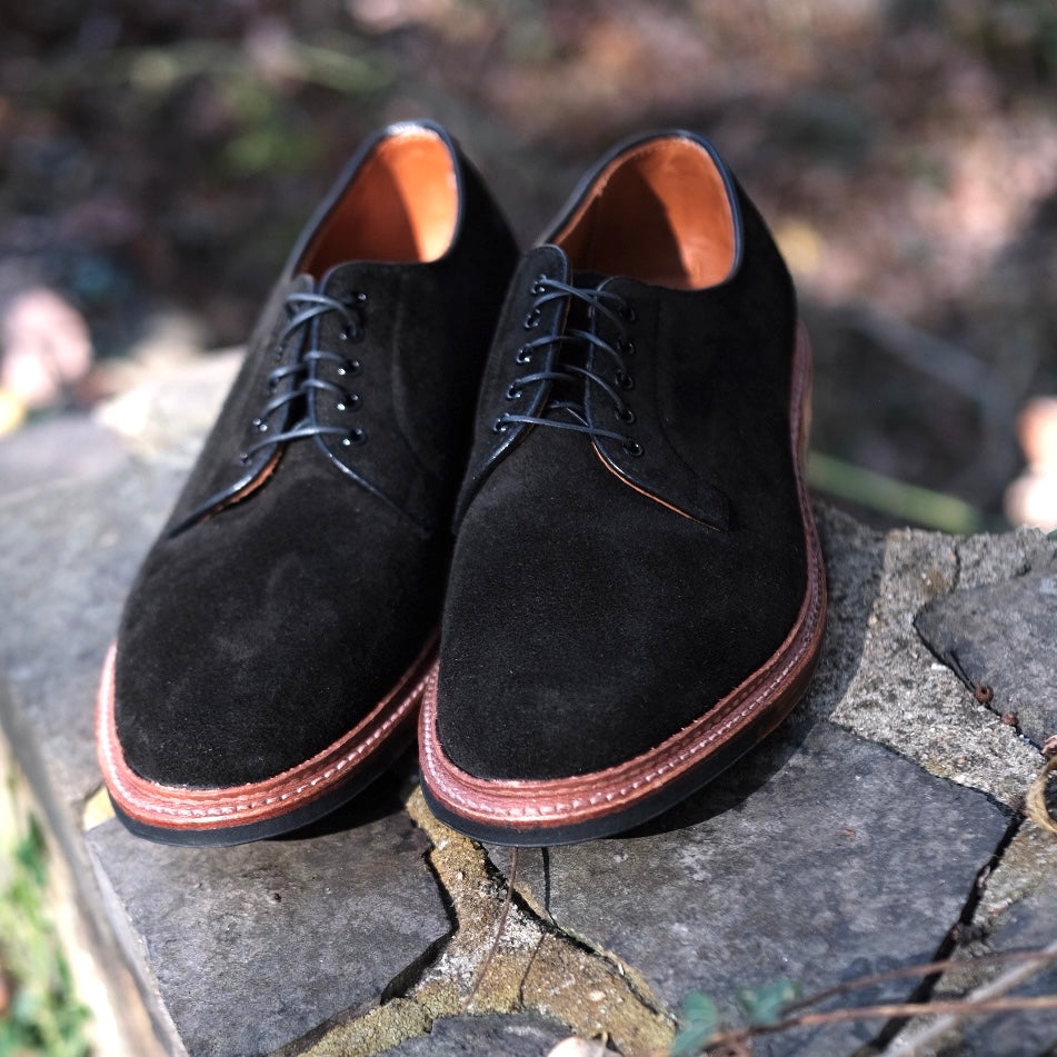 A pair of Alden Madisons D1426C - Plain Toe Blucher in Earth Reverse Chamois, featuring black laces and brown leather Barrie Last soles, rests on a stone surface outdoors, illuminated gently by sunlight.