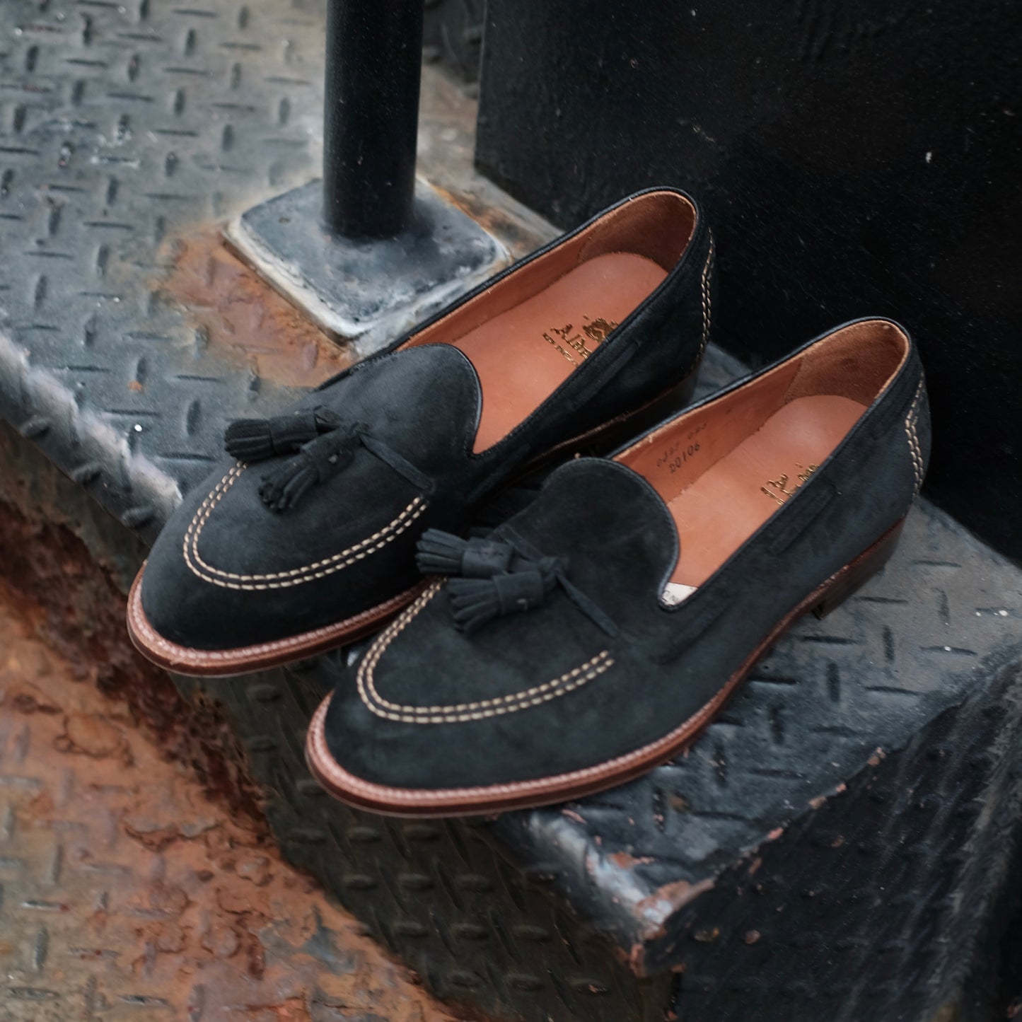 A pair of Alden Madison D0106 Black Suede Tassel Loafers with white stitching and brown soles rest on a textured metal stair.