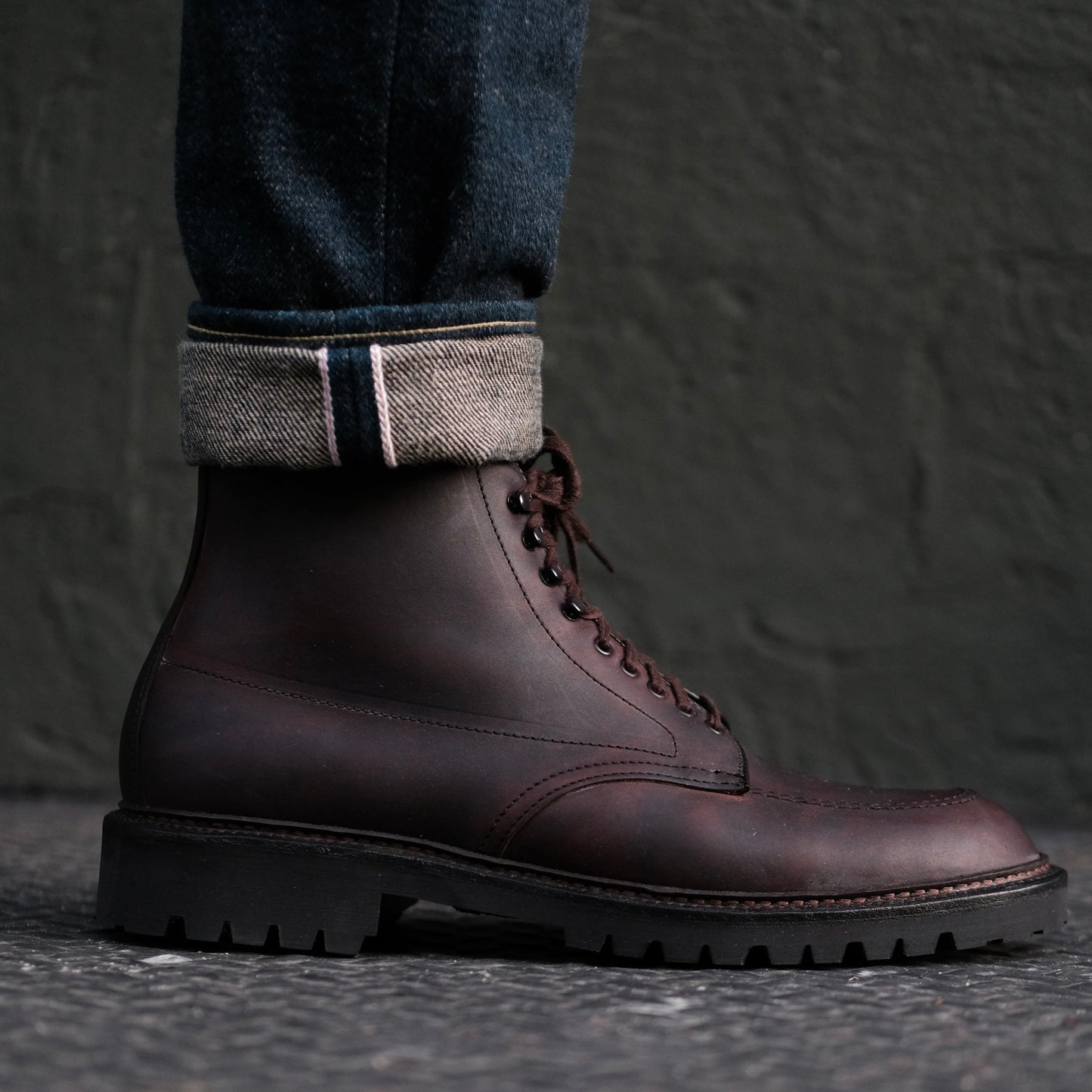 A person wears Alden Madisons 404 - Indy Boot in Kudu (Oiled CXL) with Vibram Lug Outsoles. The jeans are rolled to reveal a striped denim cuff, set against a textured dark green wall.