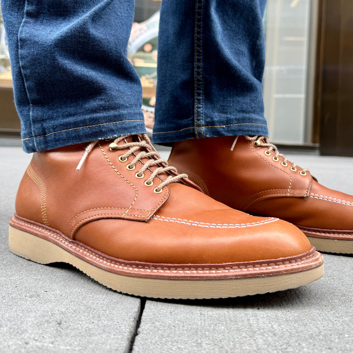 An individual wearing dark blue jeans and Alden Madisons Sherwood Indy Boot in Saddle Calf leather with cream wedge soles, stands on a textured gray surface. Designed on the Trubalance Last, these brown boots feature white stitching and are laced up.