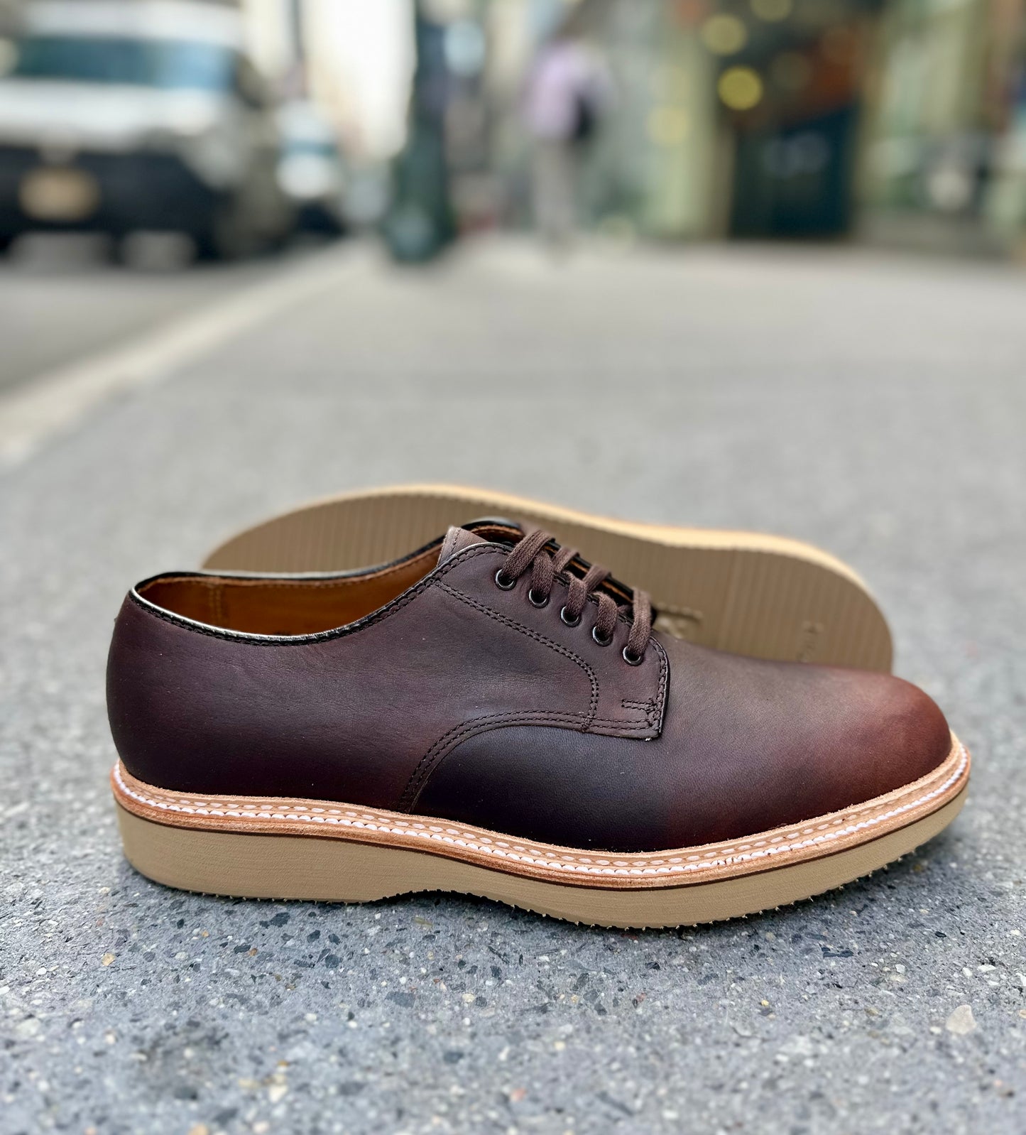 A pair of Alden Madison 29401 Plain Toe Blucher shoes in brown Kudu leather with light tan rubber soles, crafted on the Barrie last, is displayed on a city sidewalk—one upright, one on its side with the sole visible against a blurred urban background.