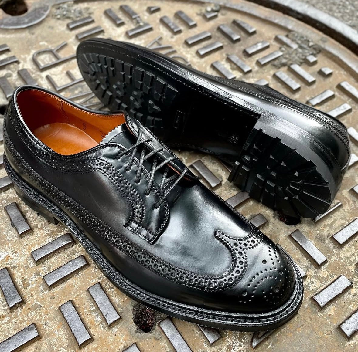 The Alden Madison 9751C Long Wing Blucher w Commando Sole in Black Shell is displayed on a metal manhole cover, highlighting its shiny leather and intricate perforated details.