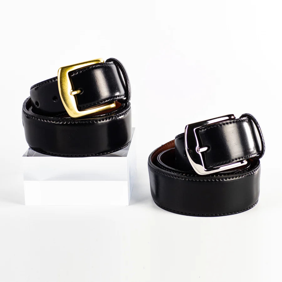 Two Alden Madison 38mm Black Shell Cordovan Belts—one with a nickel buckle, one with a brass buckle—are displayed on a white background. The brass-buckle belt is elevated on a clear acrylic block. Made in USA.