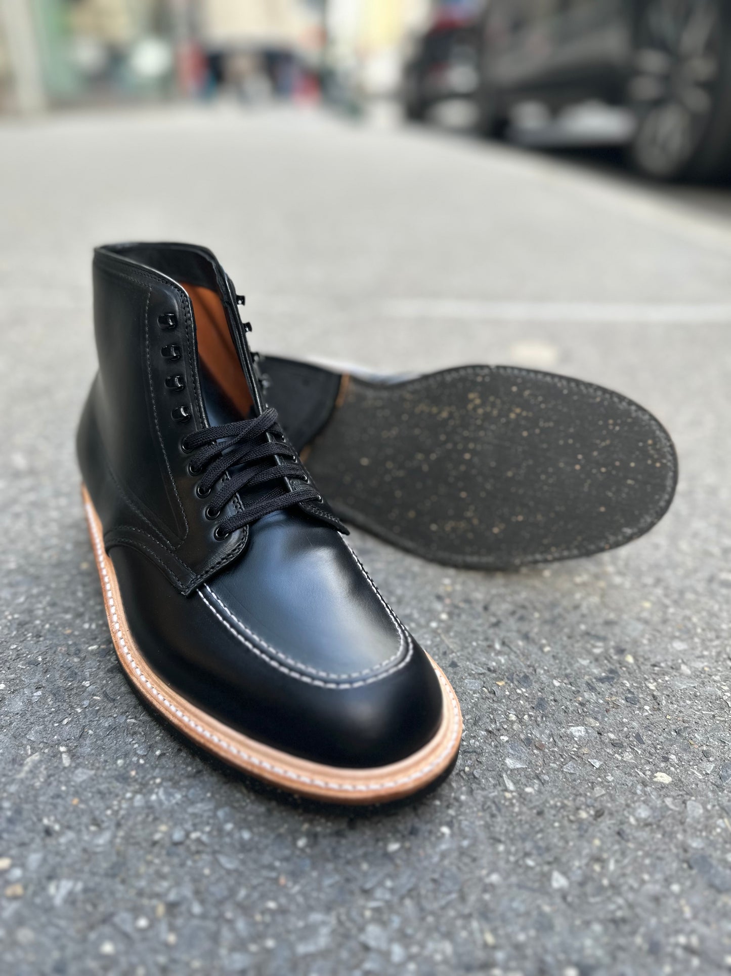 A pair of Alden Madison 401 - Indy Boot in Black Chromexcel with tan welts are shown on a paved street, one upright and one on its side to display the textured Neoprene cork sole.