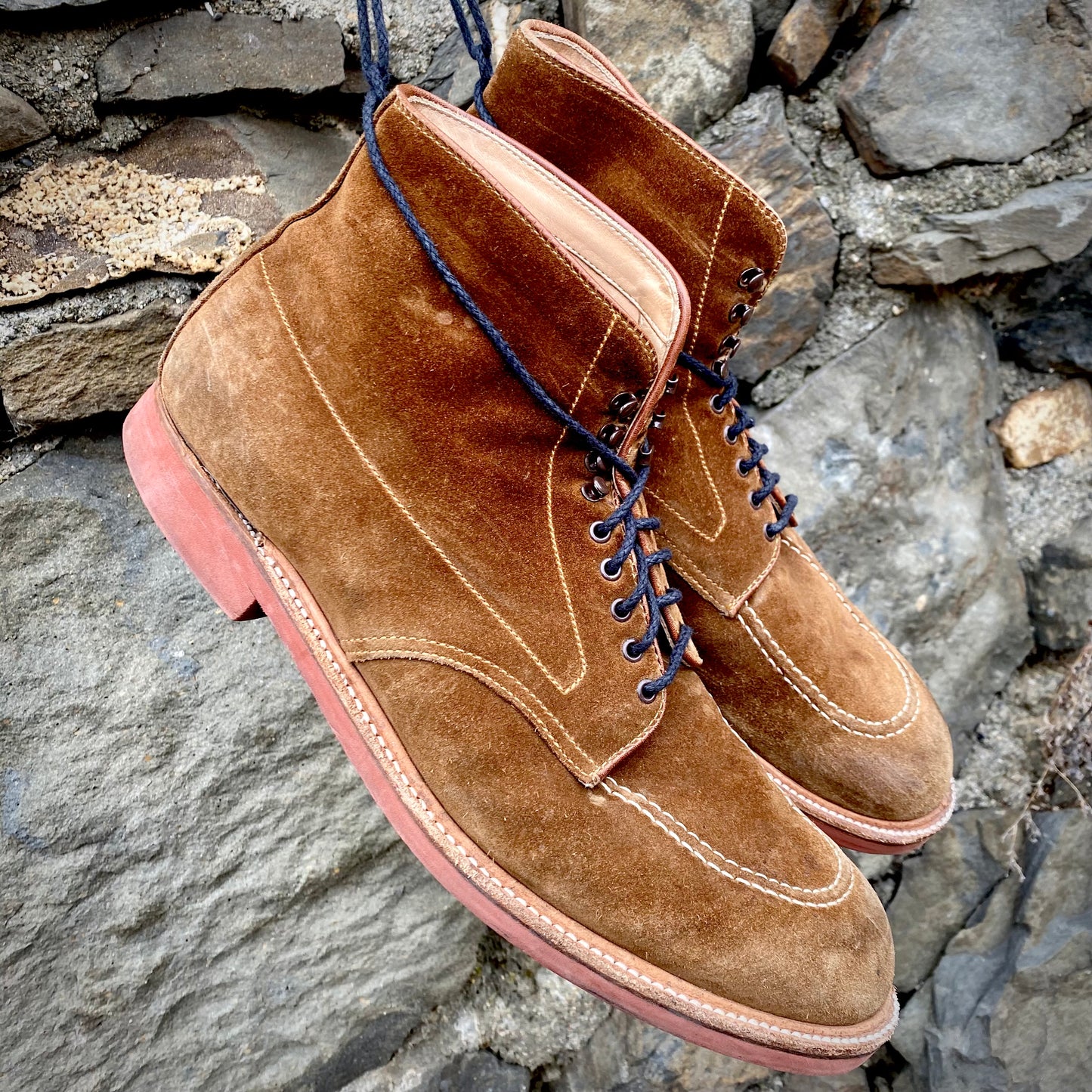 A pair of worn Alden Madisons 40531H - Redwood Indy Boot in Snuff Suede, featuring striking red rubber soles, is displayed against a rough stone wall. The boots highlight visible stitching and radiate vintage charm.