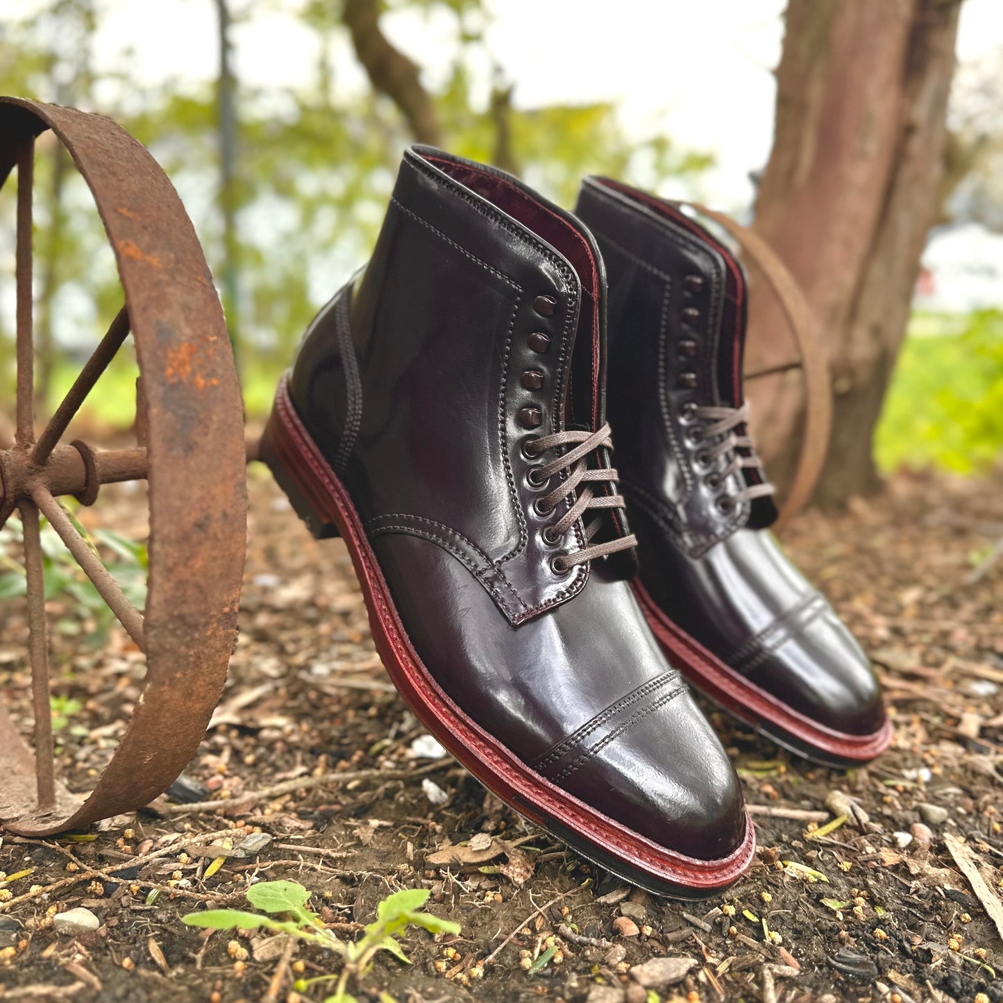 The Alden Madison D2858HC - 2/2 Cap Boot w Commando in Color 8 Shell rests outdoors on glossy dark leather with reddish-brown soles. An old rusty wheel against blurred greenery adds rustic charm to the scene.