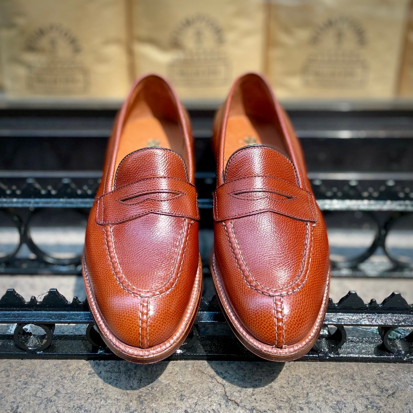 A pair of Alden Madison D1204 - Alpine Grain NST Loafers, crafted in brown leather on the Grant Last with an antique flat welt, displayed side by side on a black metal grate against a blurred tan background.