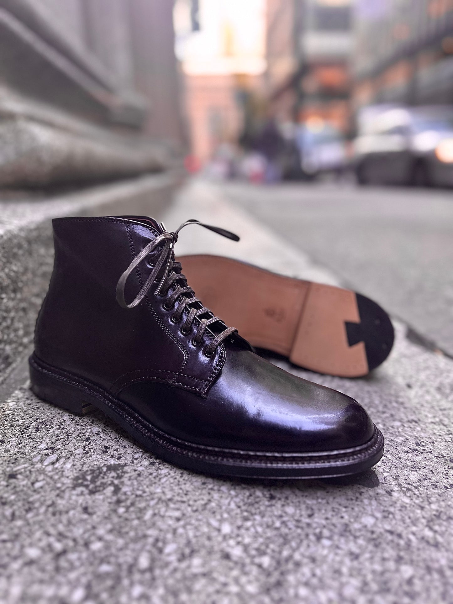 A polished Alden Madison Director boot in Color 8 Shell Cordovan sits upright on a textured stone surface, with its matching Single Oak Leather Sole beside it, set against an urban backdrop of blurred buildings and passing cars.