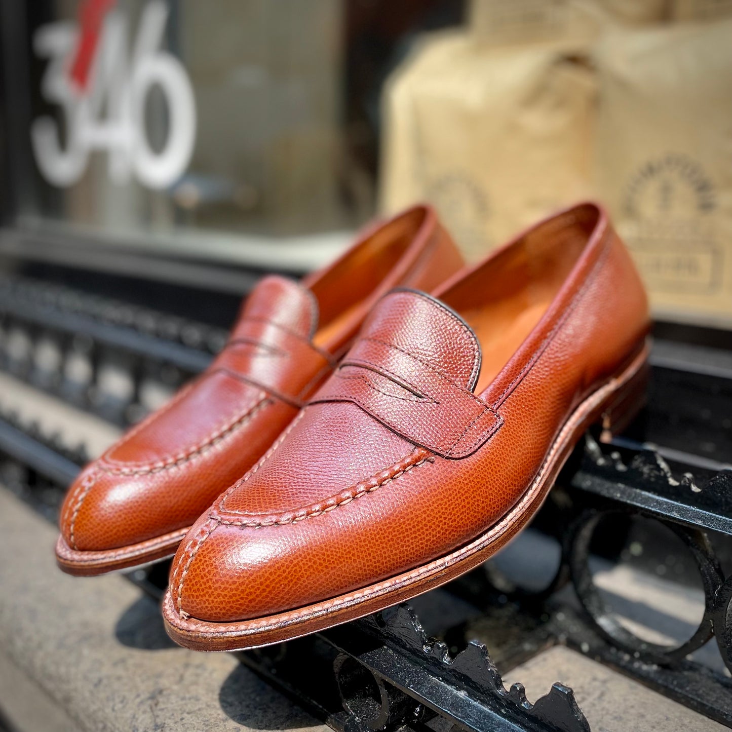 The Alden Madison D1204 - Alpine Grain NST Loafer, crafted from brown alpine grain leather on the Grant Last with an antique flat welt, is displayed outdoors on a black metal railing; blurred in the background are the number 346 and tan bags.