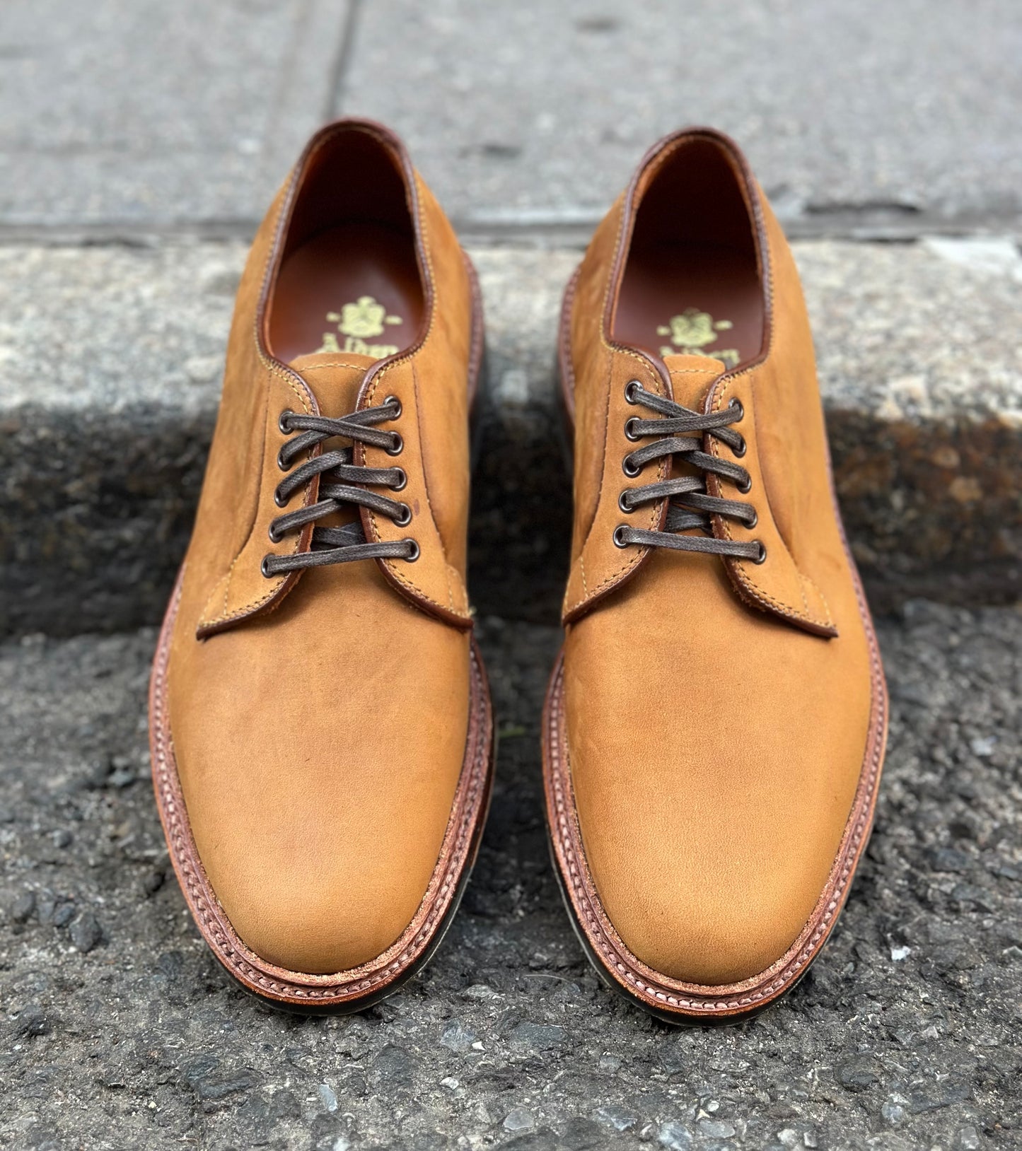 A pair of Alden Madison 9475C Plain Toe Blucher shoes in tan smooth chamois, featuring a Commando sole and brown laces, sits side by side on a rough concrete surface near the curb.