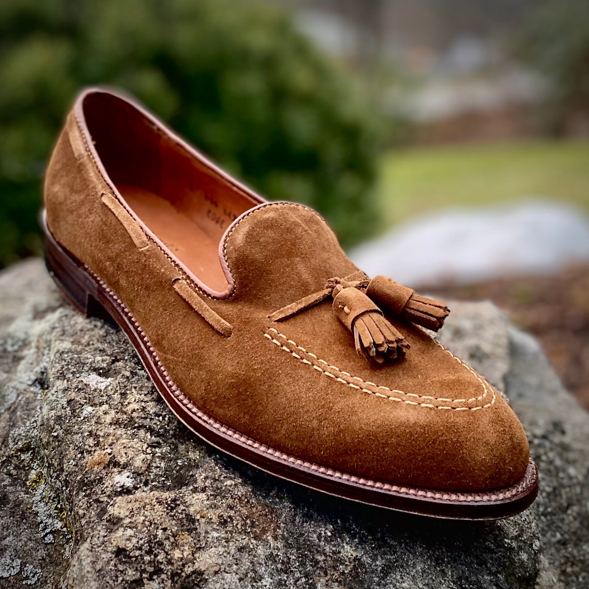 The Alden Madison 3403 Tassel Loafer in Snuff Suede, featuring detailed stitching and twin tassels on the upper, rests on a rock with a leather sole. The backdrop is blurred greenery and rocks, emphasizing its classic Aberdeen Last silhouette.