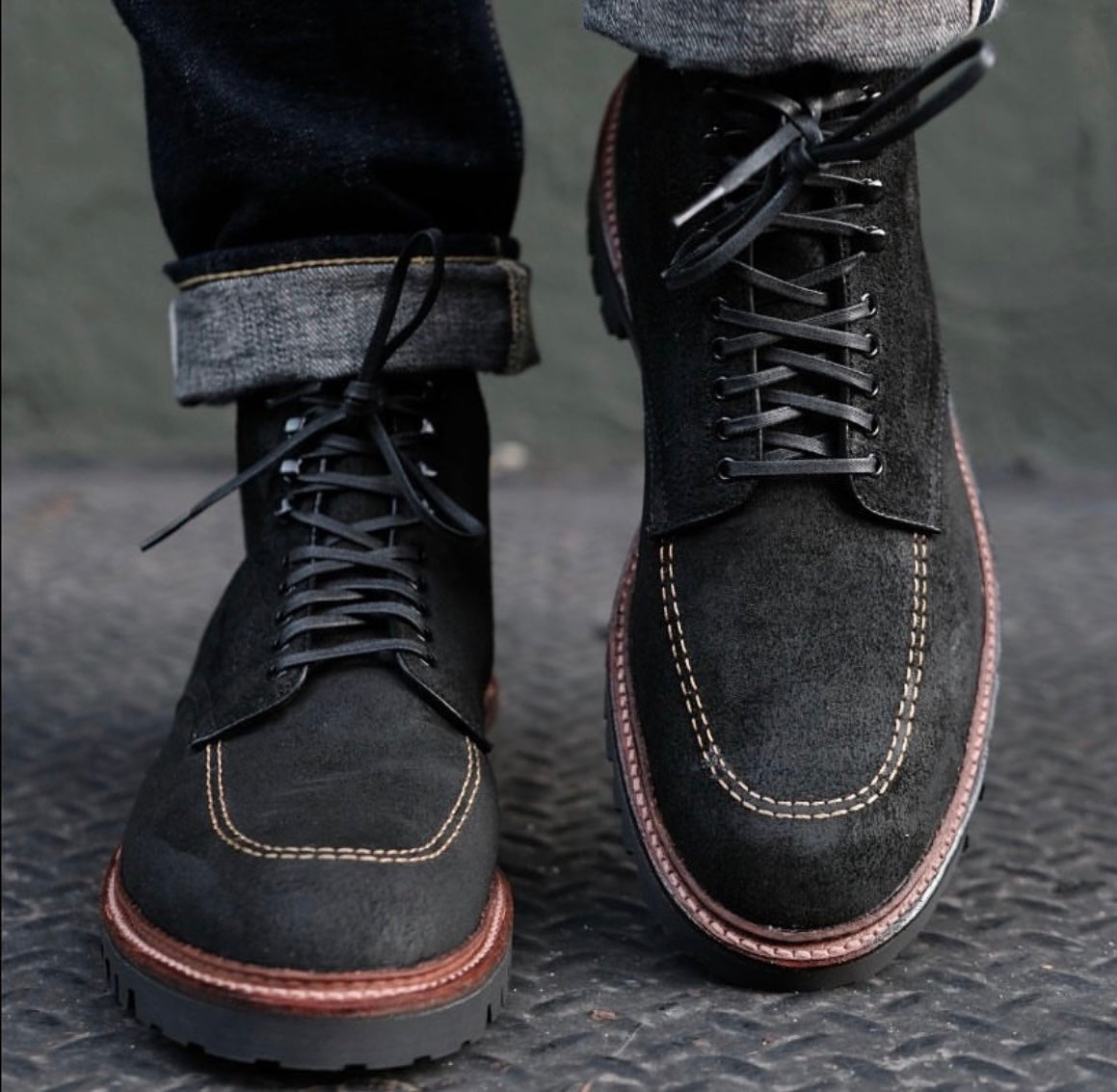 A close-up of a person wearing cuffed jeans and Alden Madison’s D9916H - Indy Boot in Earth Reverse Chamois, featuring black suede, contrast stitching, brown soles, and a Vibram Heavy Lug Outsole on a textured metal surface.
