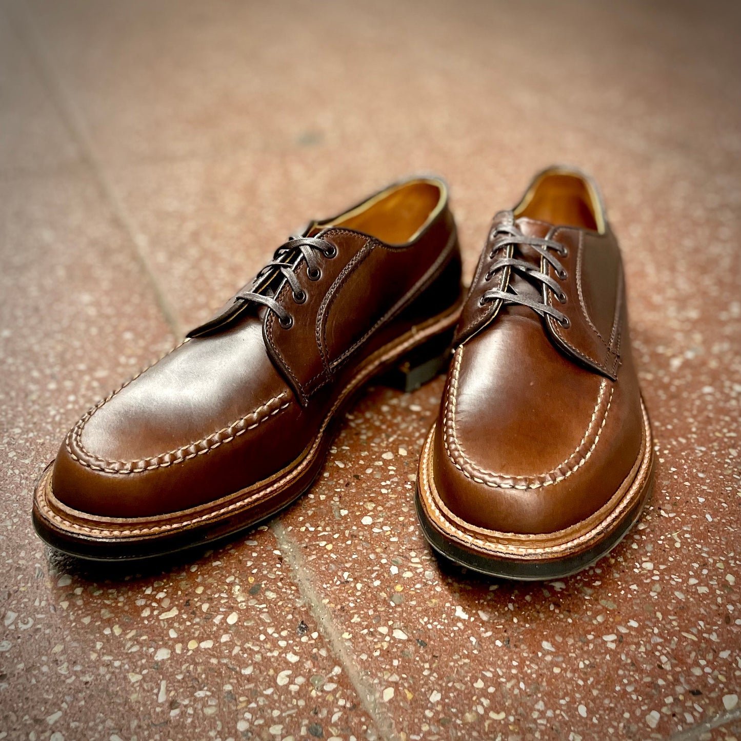 A pair of Alden Madison D1611C - Ranger Mocc in Brown CXL shoes features exquisite stitching around the toes. Positioned on a speckled, reddish-brown tiled floor, they cast soft shadows.