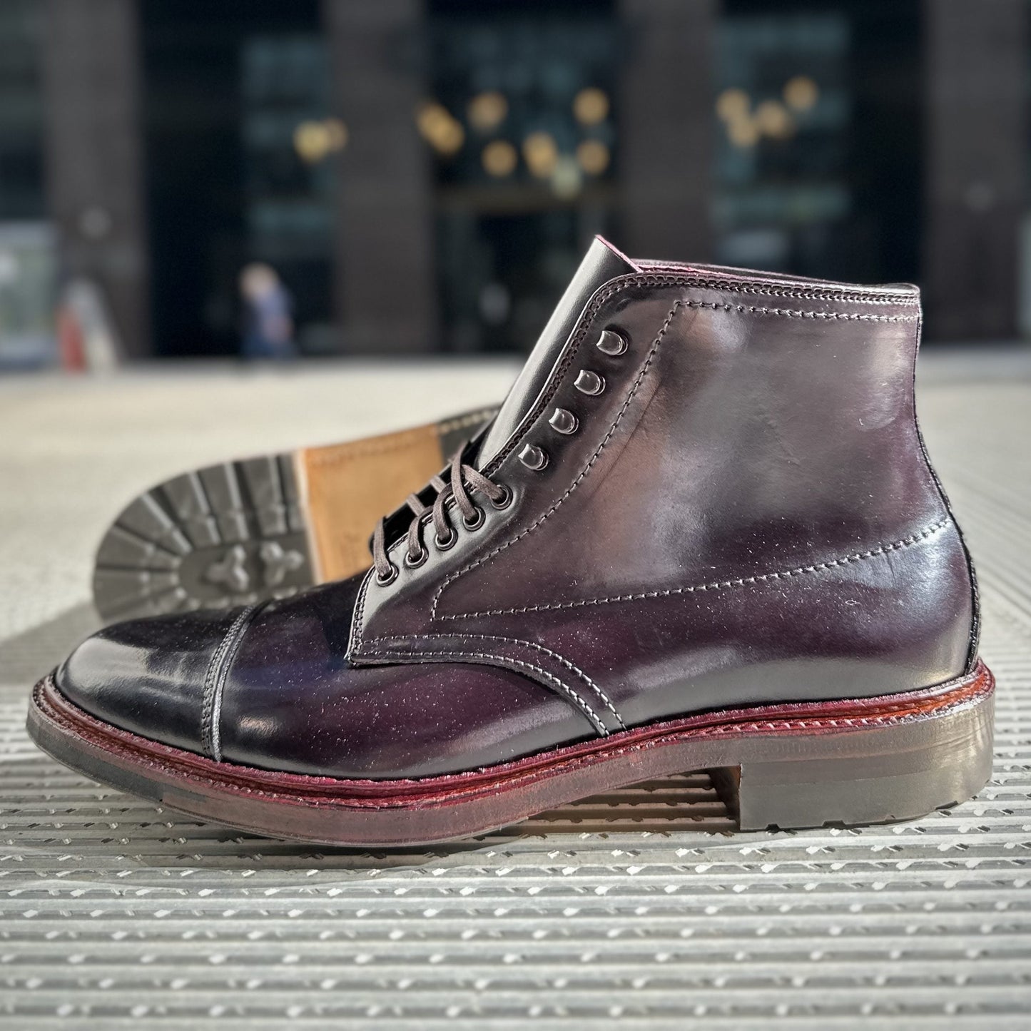 An Alden Madison D2824HC Cap Toe Boot in Color 8 Shell with a Commando Sole features red stitching and untied laces, resting on a textured surface with a blurred building façade in the background.